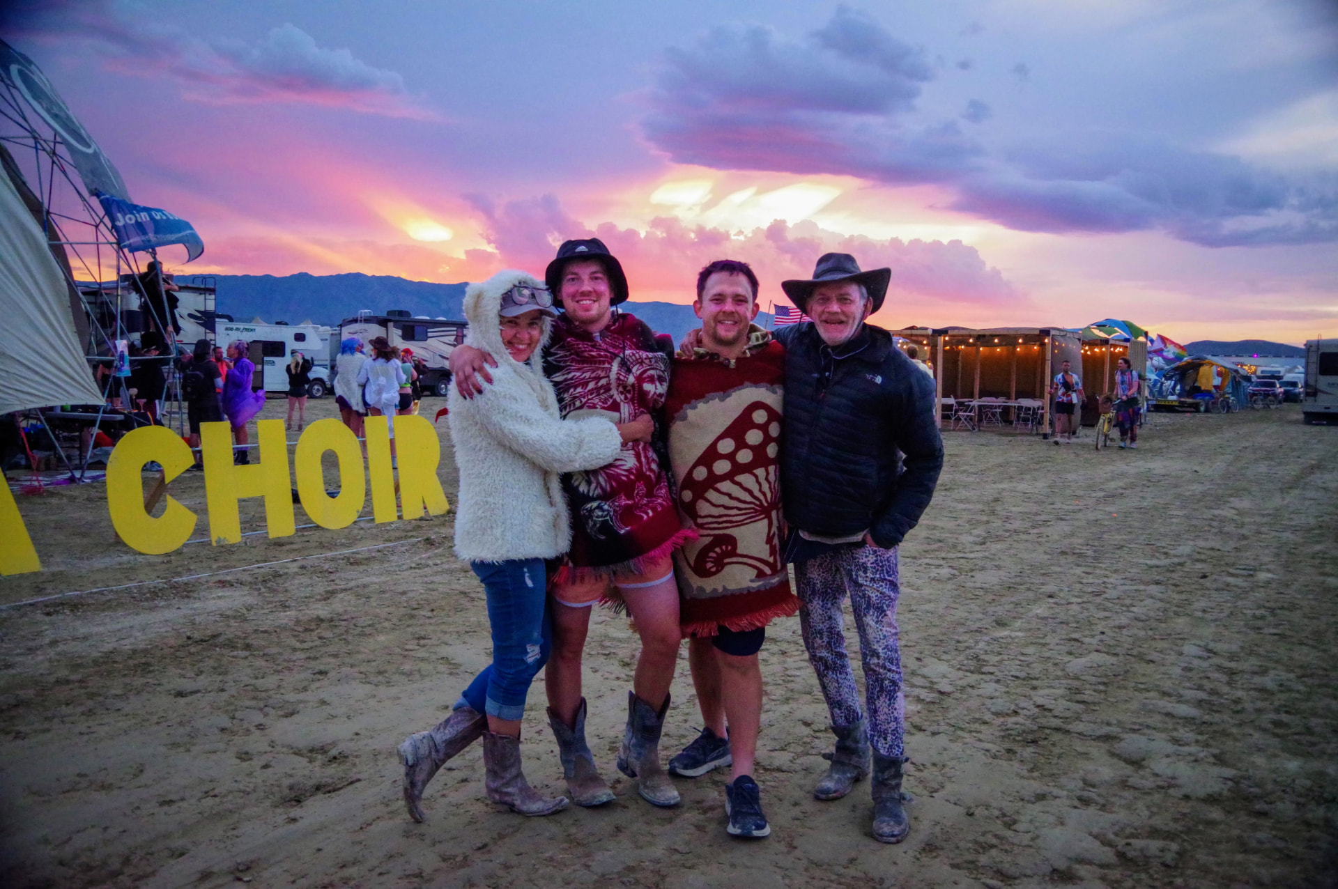 Four campers in blankets and coats at sunset by the CHOIR sign